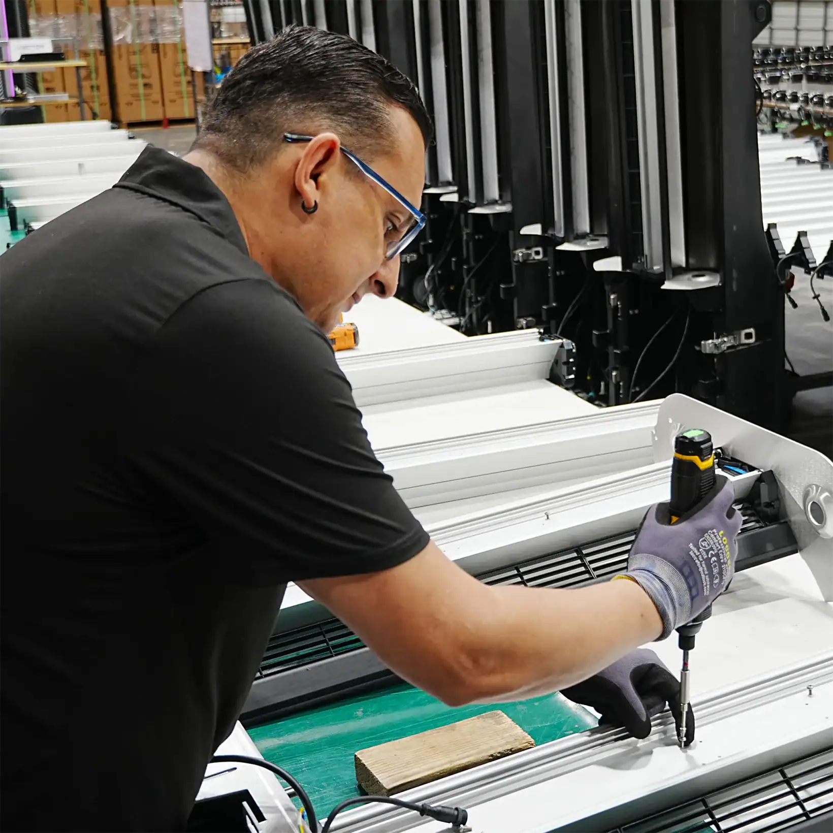 A worker in safety gear uses a power tool to assemble metal frames in a factory.