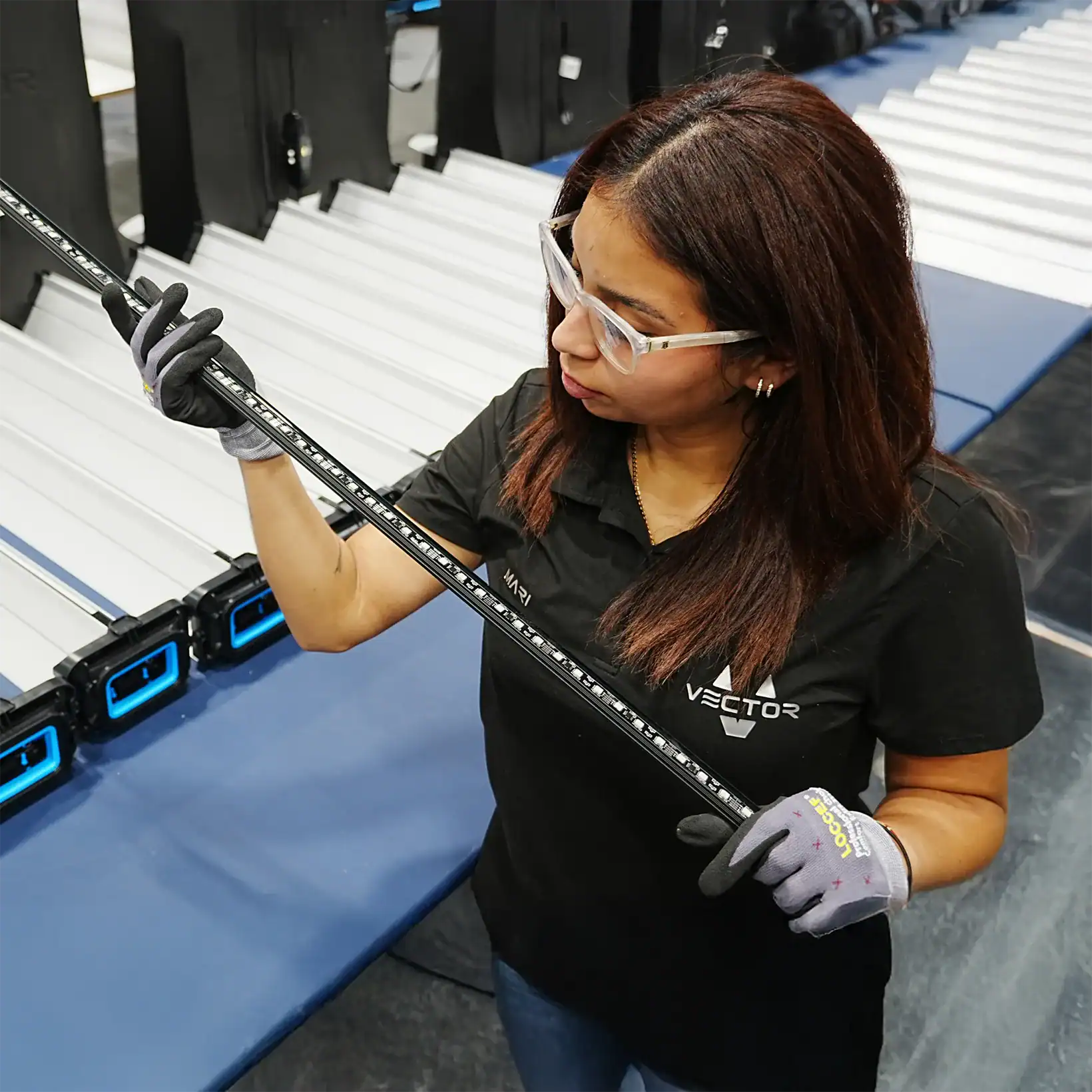 A woman in safety glasses and gloves inspects a long, slim electronic device in a tech facility.