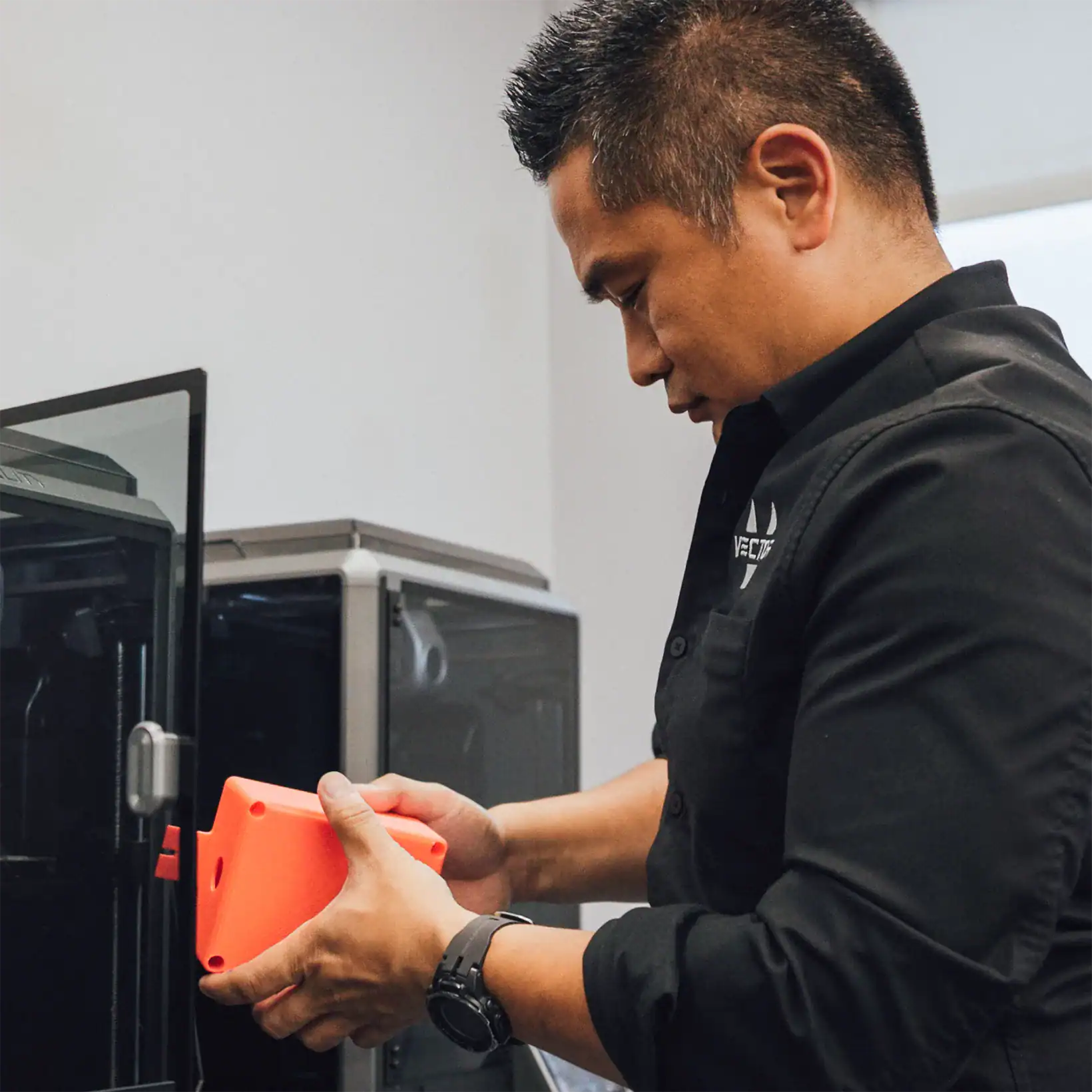 A man in a black shirt examines a bright red 3D printed object beside a 3D printer, displaying focus and precision in a modern workshop setting.