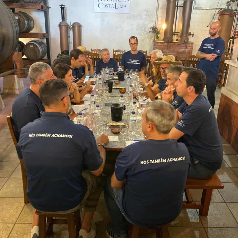 Degustação de vinhos na Vinícola Venâncio da Costa Lima, com participantes sentados ao redor de uma mesa, vestindo camisolas com a mensagem "NÓS TAMBÉM ACHAMOS!", em ambiente rústico com barris de vinho ao fundo.