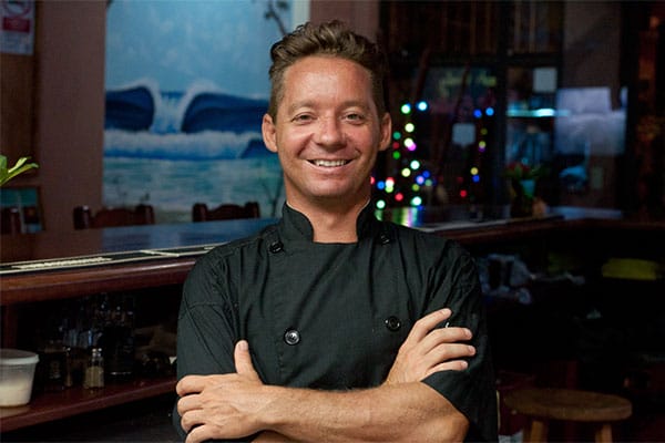 Chef in restaurant smiling with arms crossed in front of a bar area, festive decorations in the background.