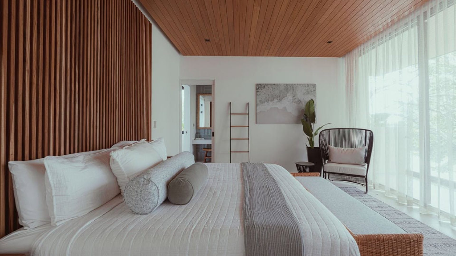 Modern bedroom with wooden ceiling and natural light, featuring minimalist decor and cozy seating area.