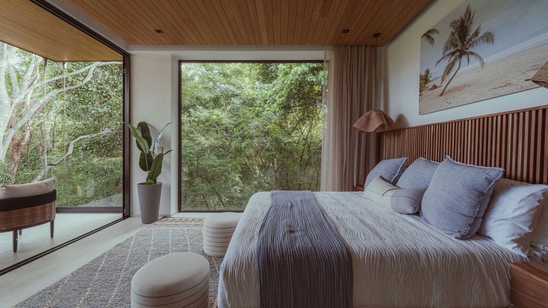 Luxury bedroom with large glass windows overlooking lush greenery and a tropical beach scene, modern and cozy interior design, Vida Pinilla.