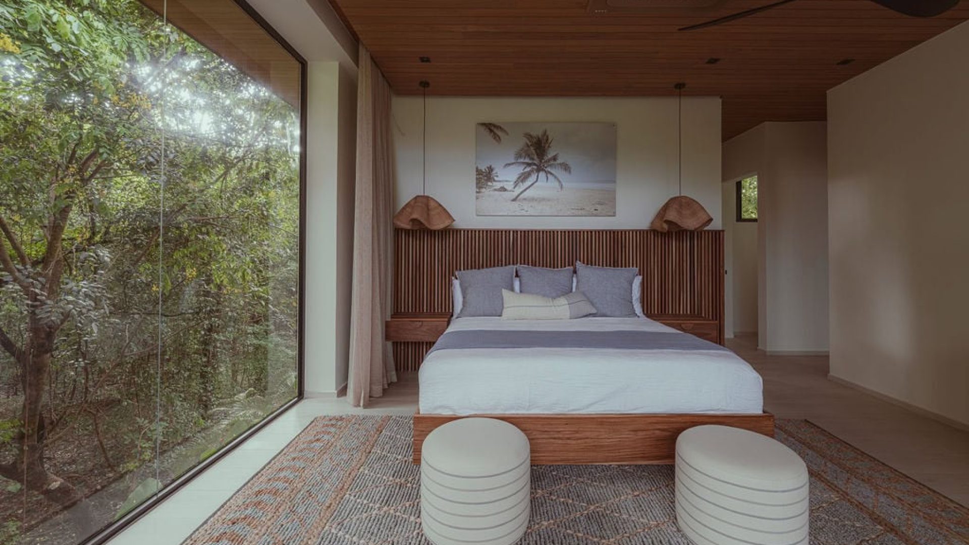 Serene bedroom with large floor-to-ceiling window overlooking lush greenery, modern decor, and beach-themed artwork.