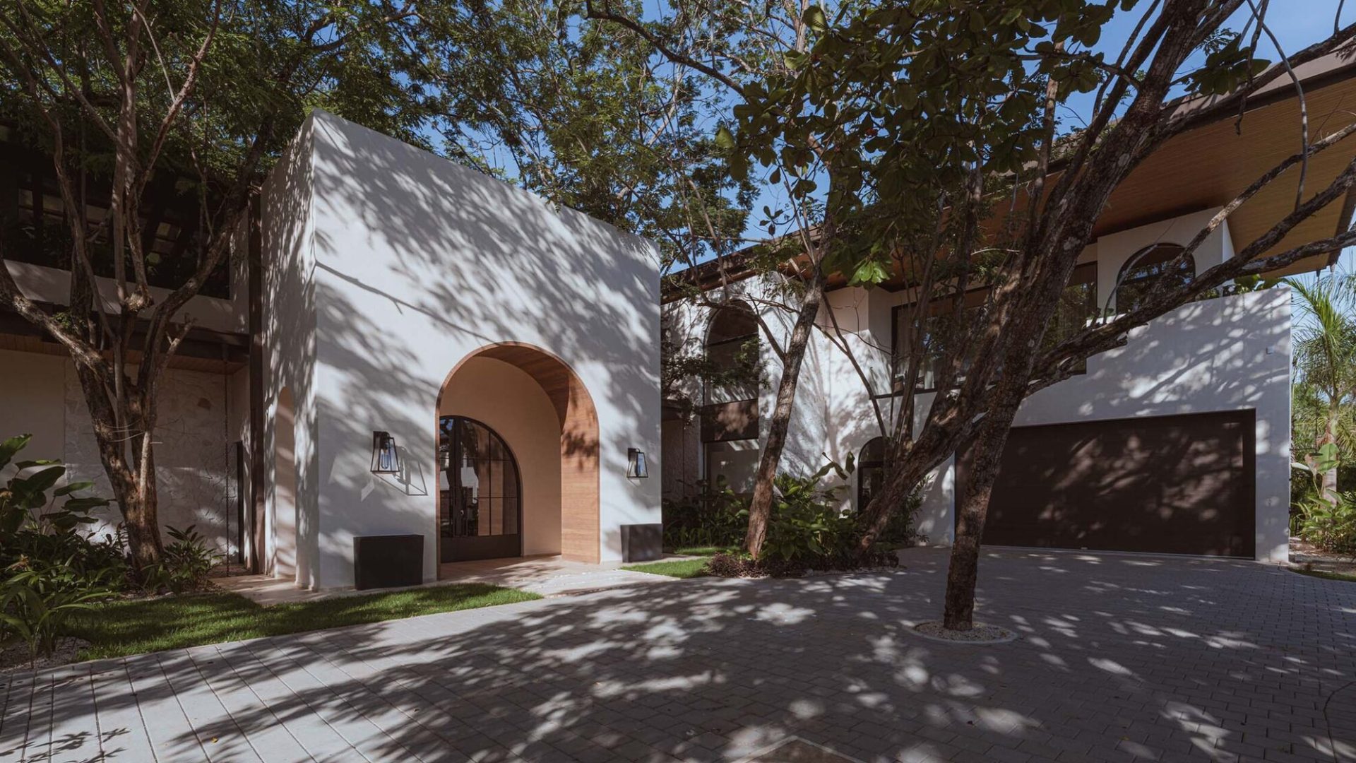Modern luxury house with sleek white exterior, arched entryway, and lush tree shadows for a tropical vibe.