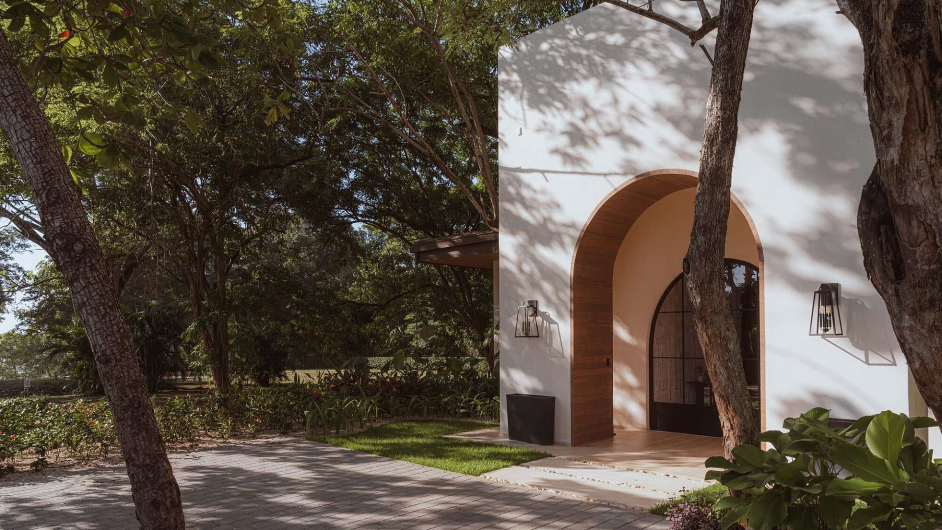 Modern house with white walls and wooden arch door surrounded by lush trees.