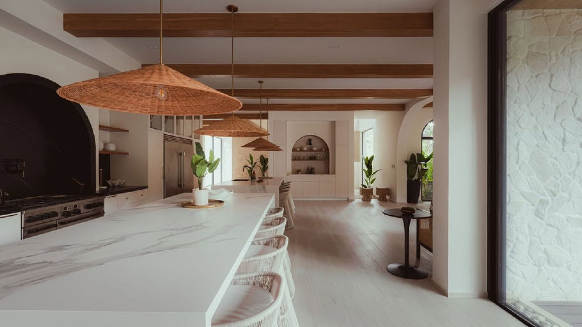 Modern minimalist kitchen with white marble island and wooden ceiling beams, natural light, indoor plants, and contemporary decor.