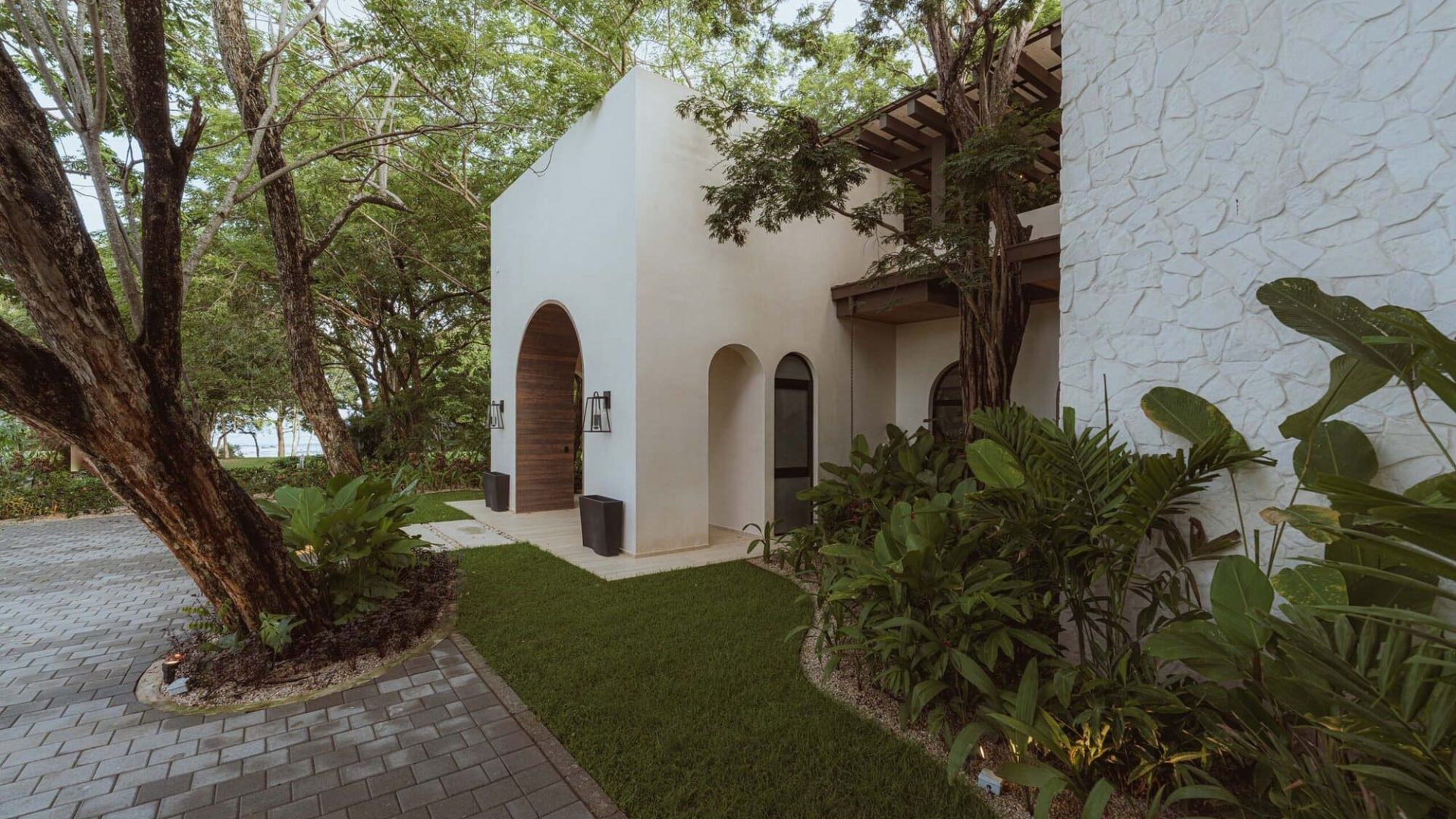 Modern villa with lush greenery, white stucco walls, and arched wooden doorways in a tropical setting.