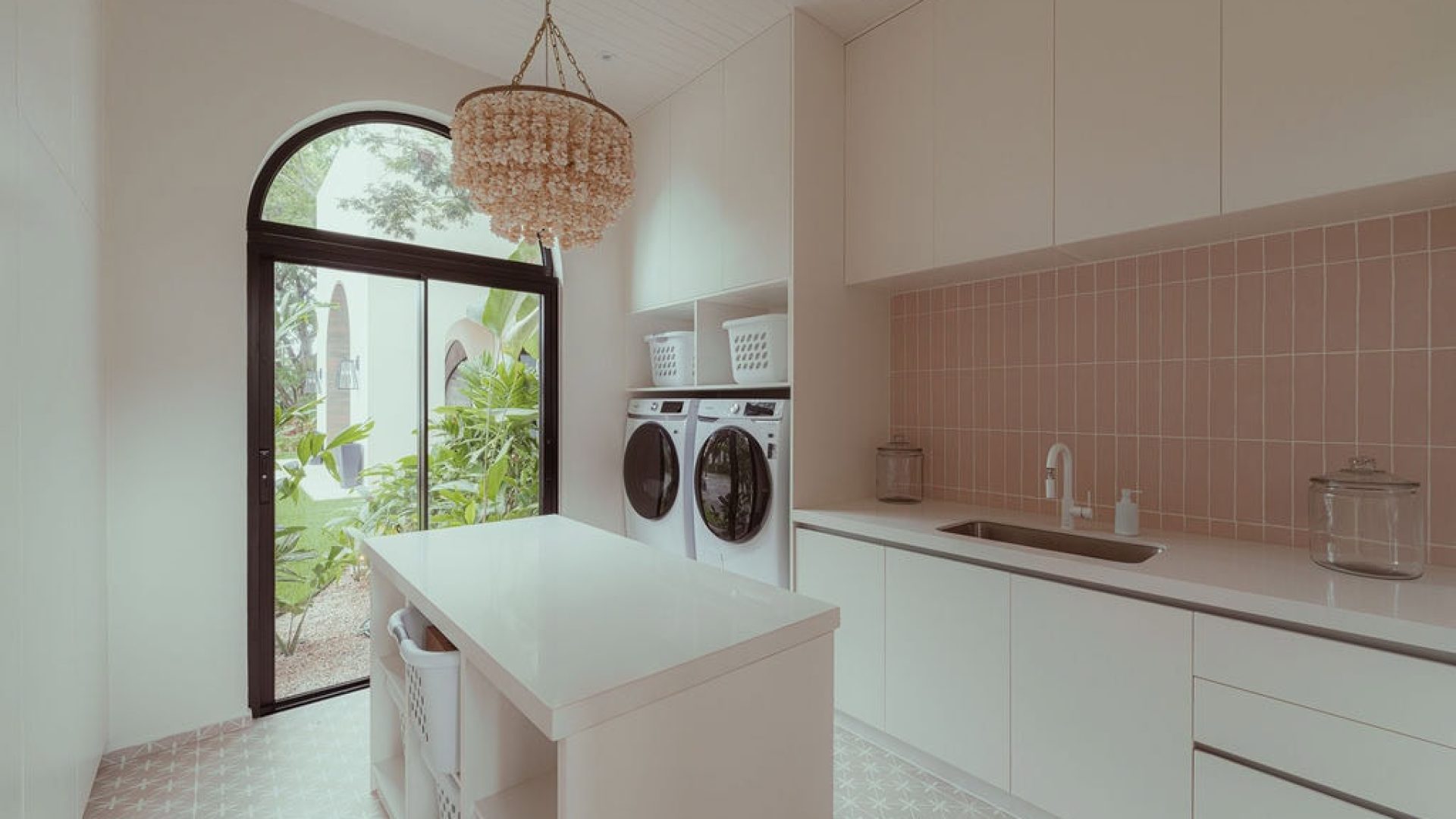 Bright laundry room with white cabinets, pink tiled backsplash, and large window overlooking garden.