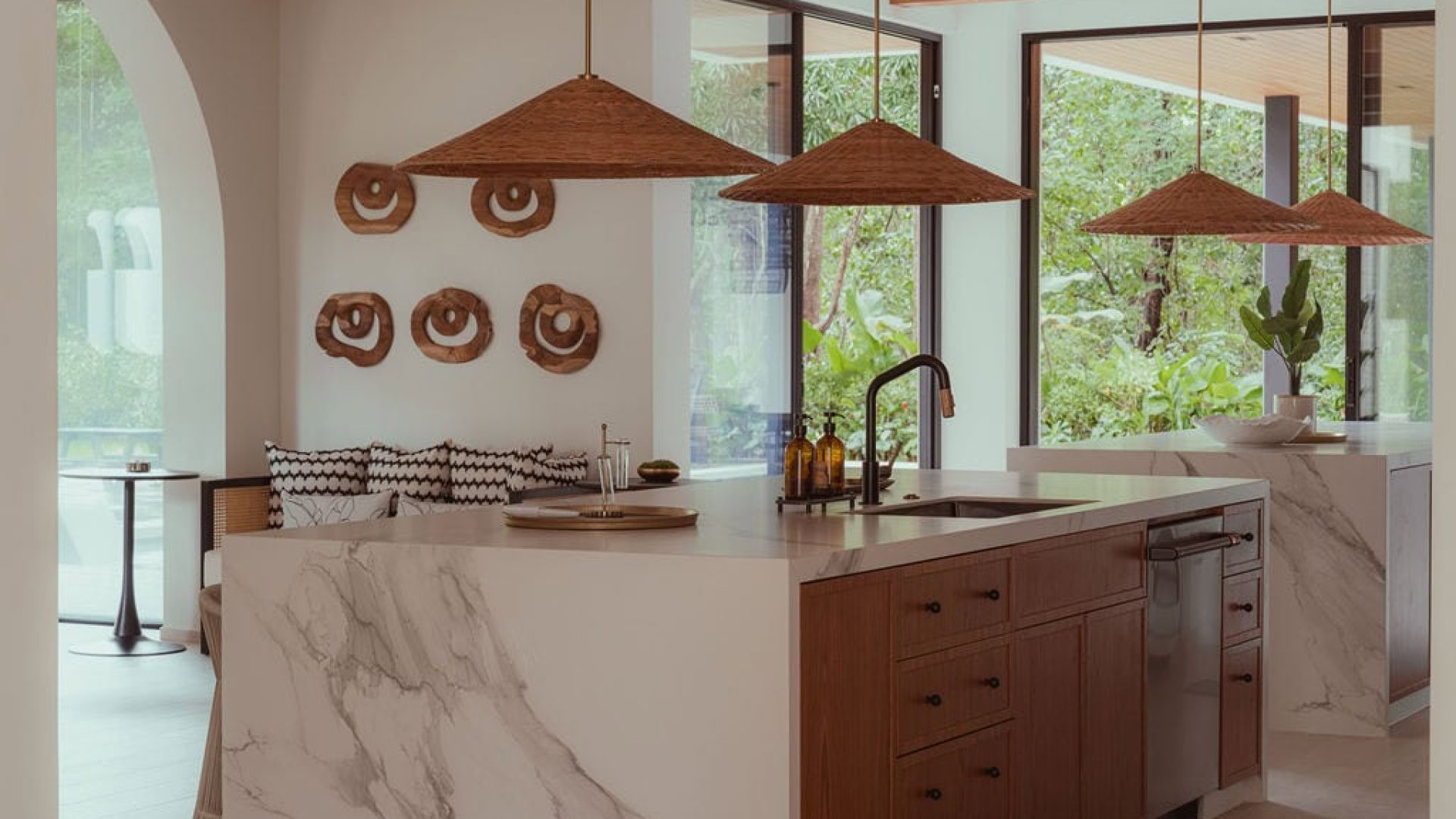Contemporary kitchen with marble island, wooden cabinets, and large windows showcasing greenery.