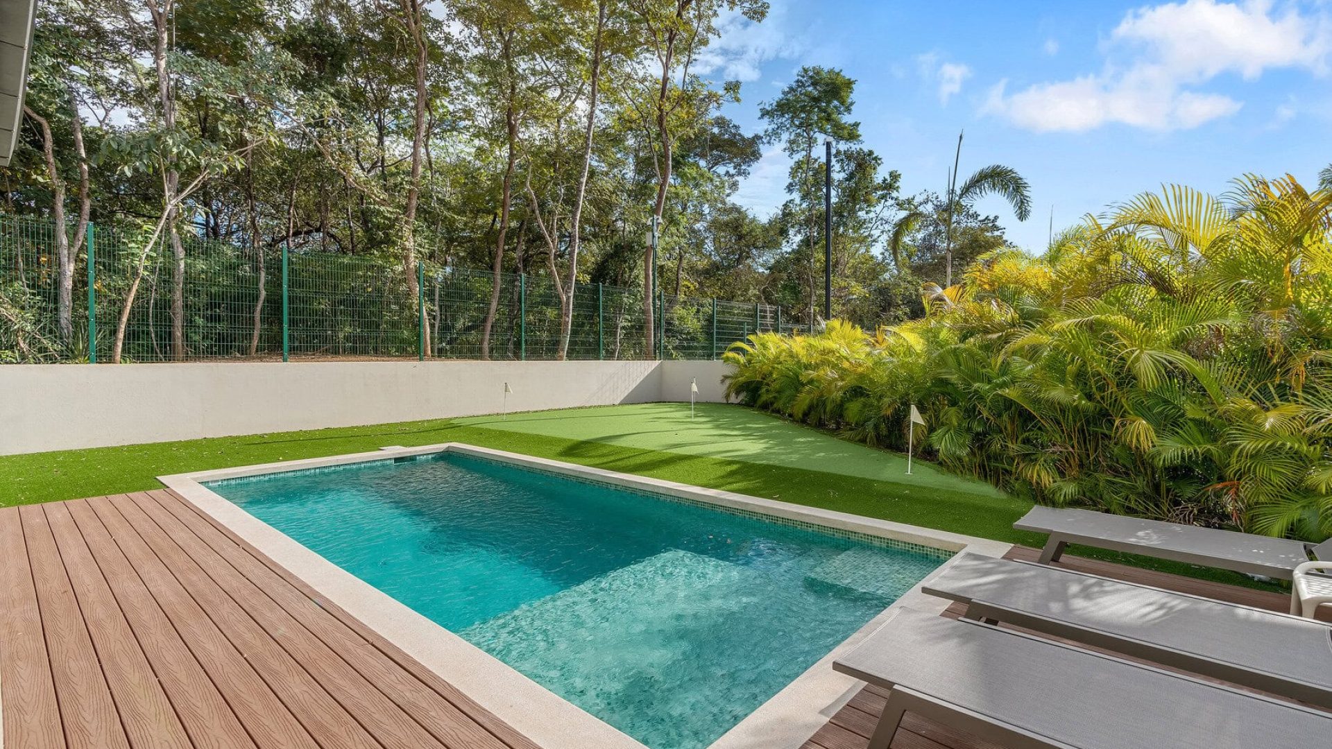 Lush backyard swimming pool with wooden deck and tropical plants, perfect for relaxation.