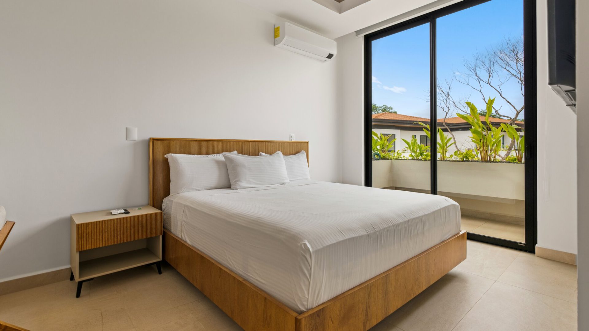 Bright modern hotel bedroom with wooden furniture and balcony view of tropical plants and clear sky.