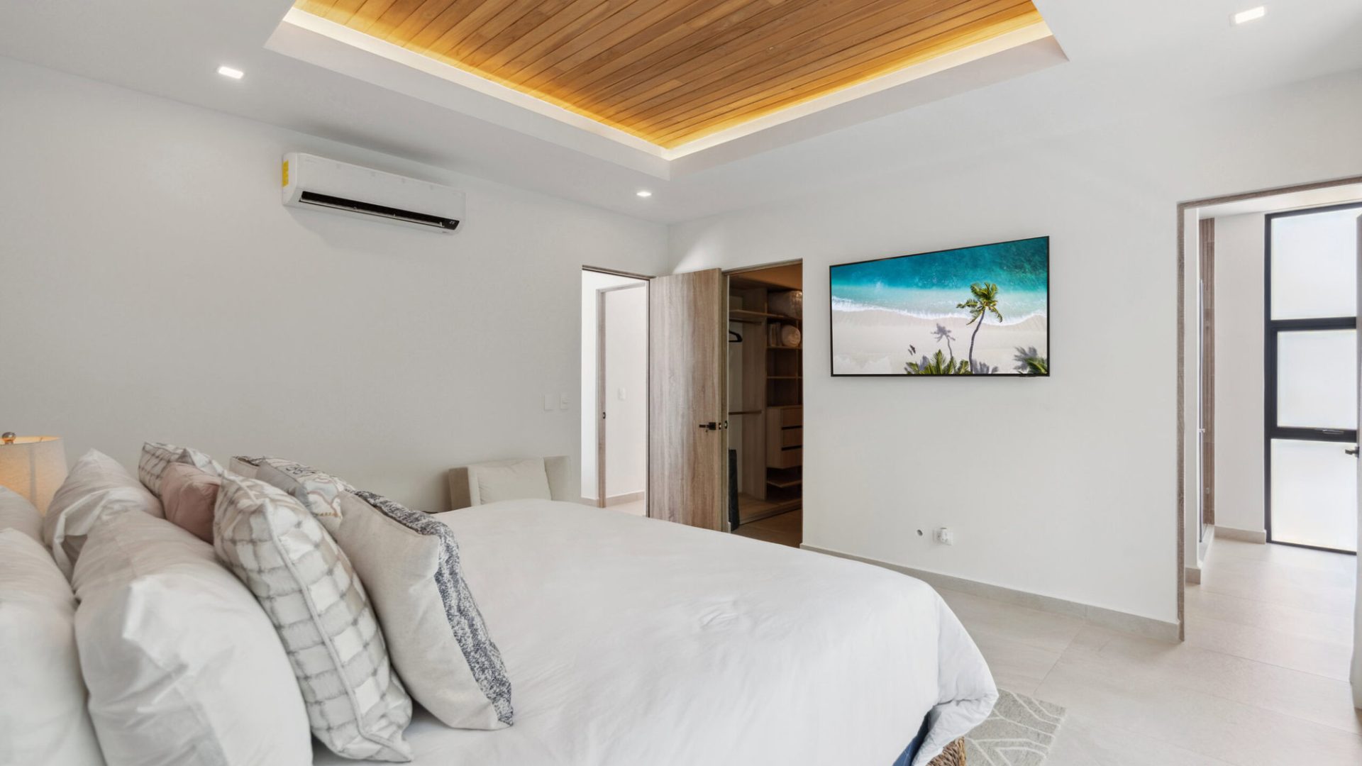 Modern bedroom with white walls, wooden ceiling accents, and tropical beach art on screen.