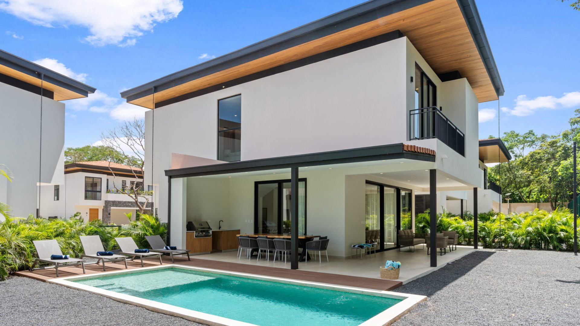 Modern luxury house with pool, patio, and outdoor dining area in a lush green setting.