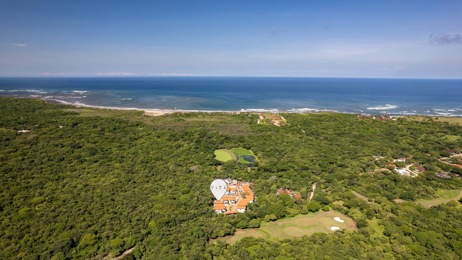 Sea view of lush green forest, ocean coastline, and Vida Pinilla housing community with golf course, aerial drone shot.
