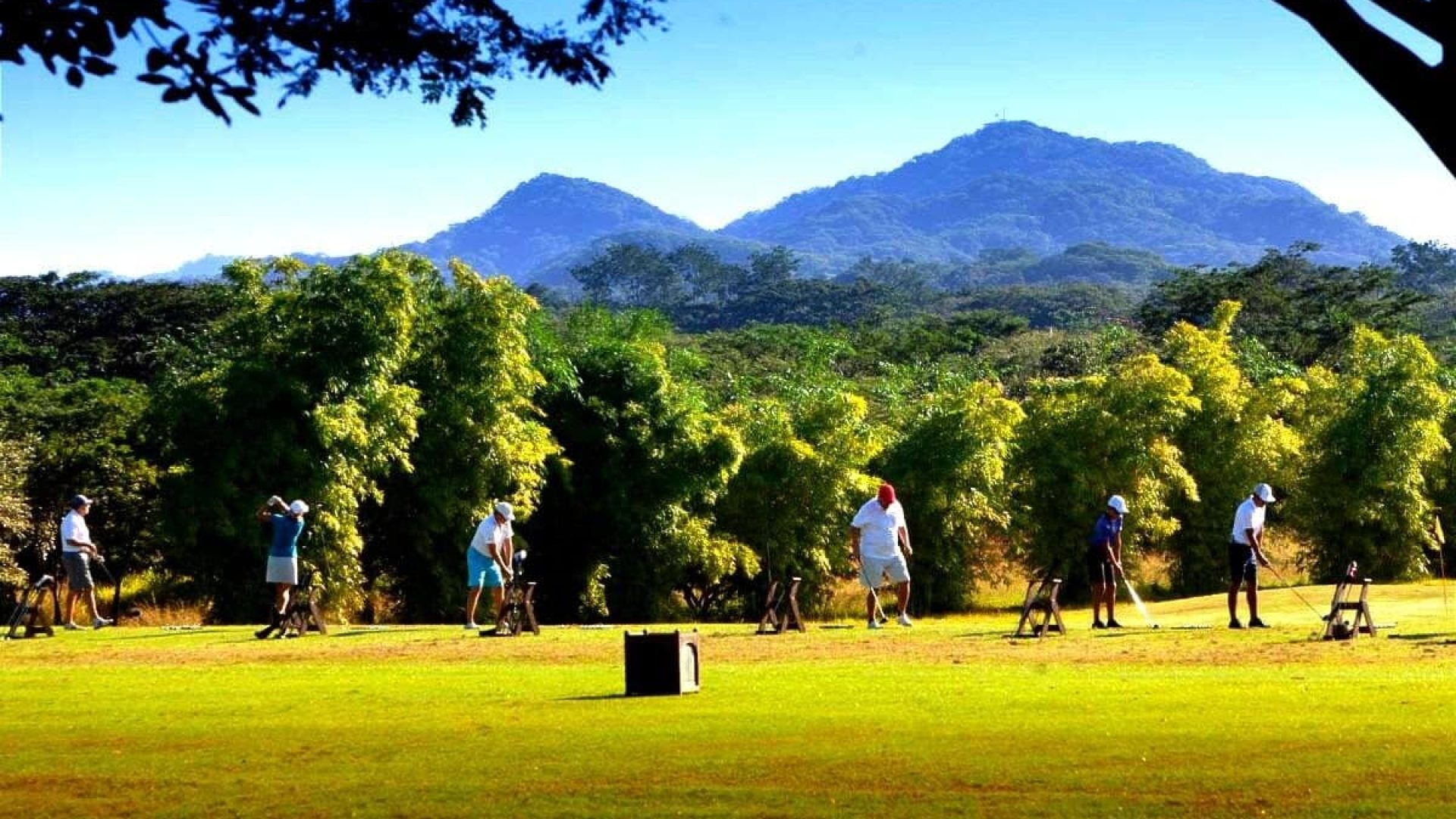 Golf players practicing on a lush green course with mountains and trees in the background.