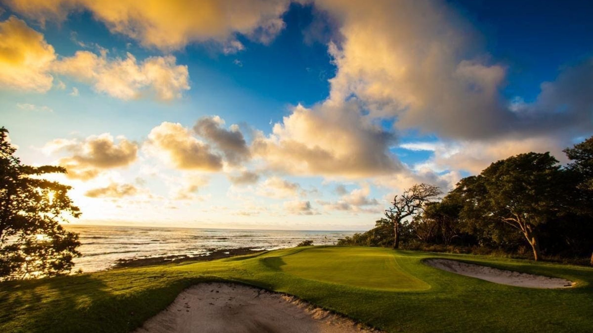 Scenic golf course by the ocean during sunset with cloud-filled sky and lush green landscape.