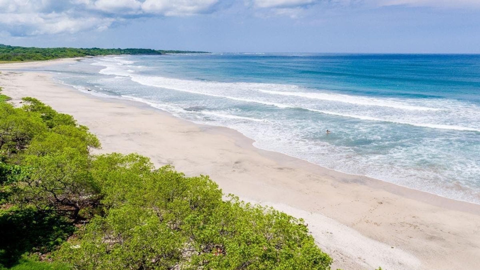 Scenic beach with white sand, clear blue ocean, and lush green trees under a partly cloudy sky.