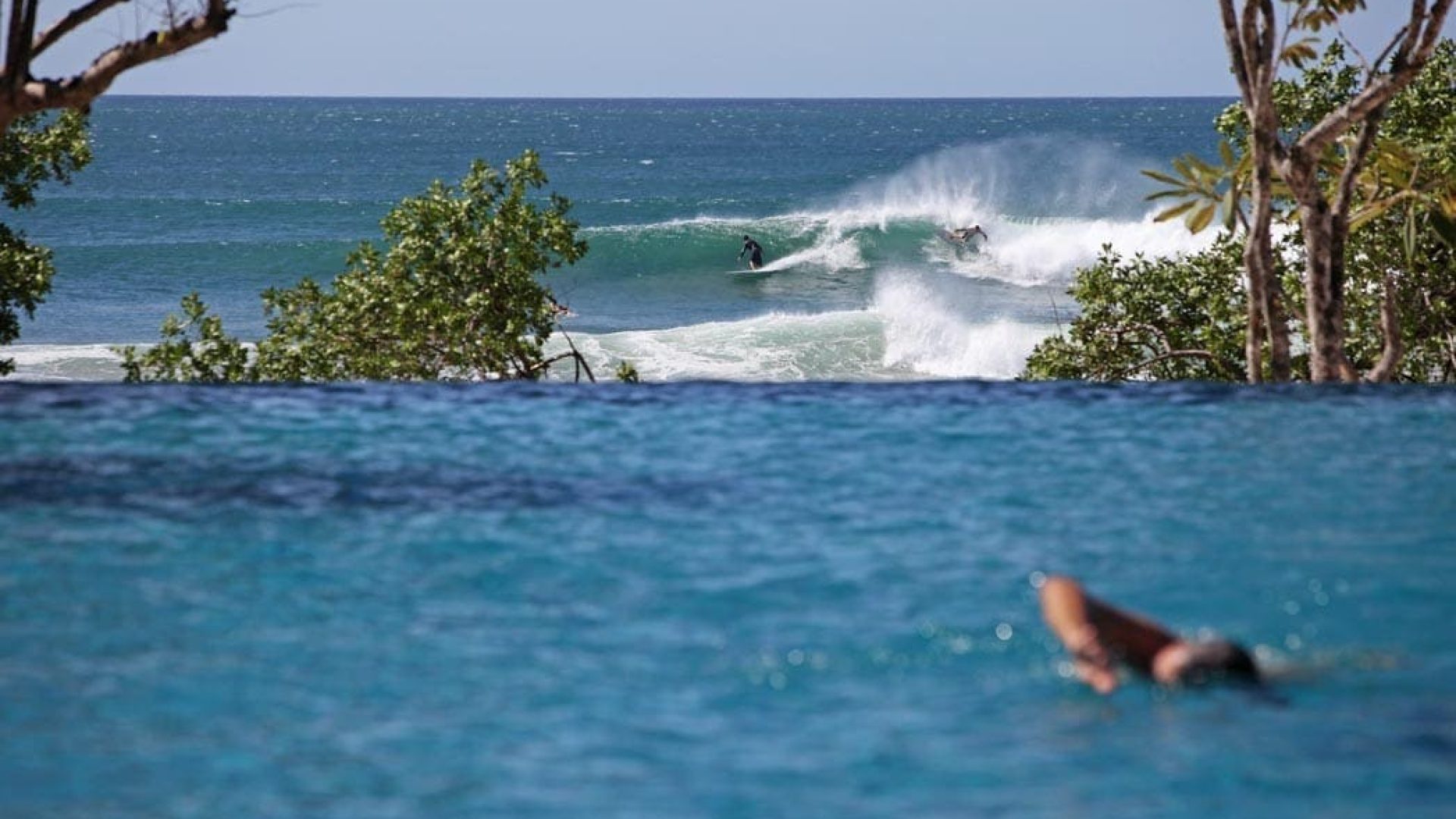 Sunset over ocean with surfers riding waves in a tropical beach setting.