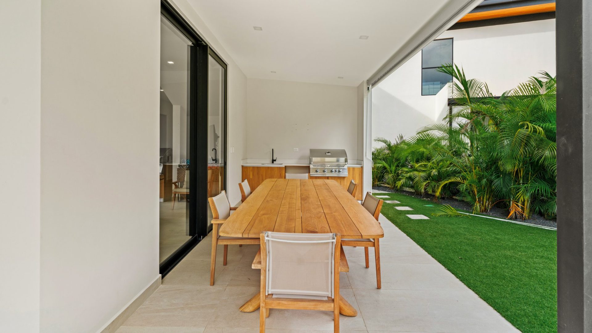 Modern outdoor dining area with wooden table, chairs, and built-in grill on a terrace with lush greenery.