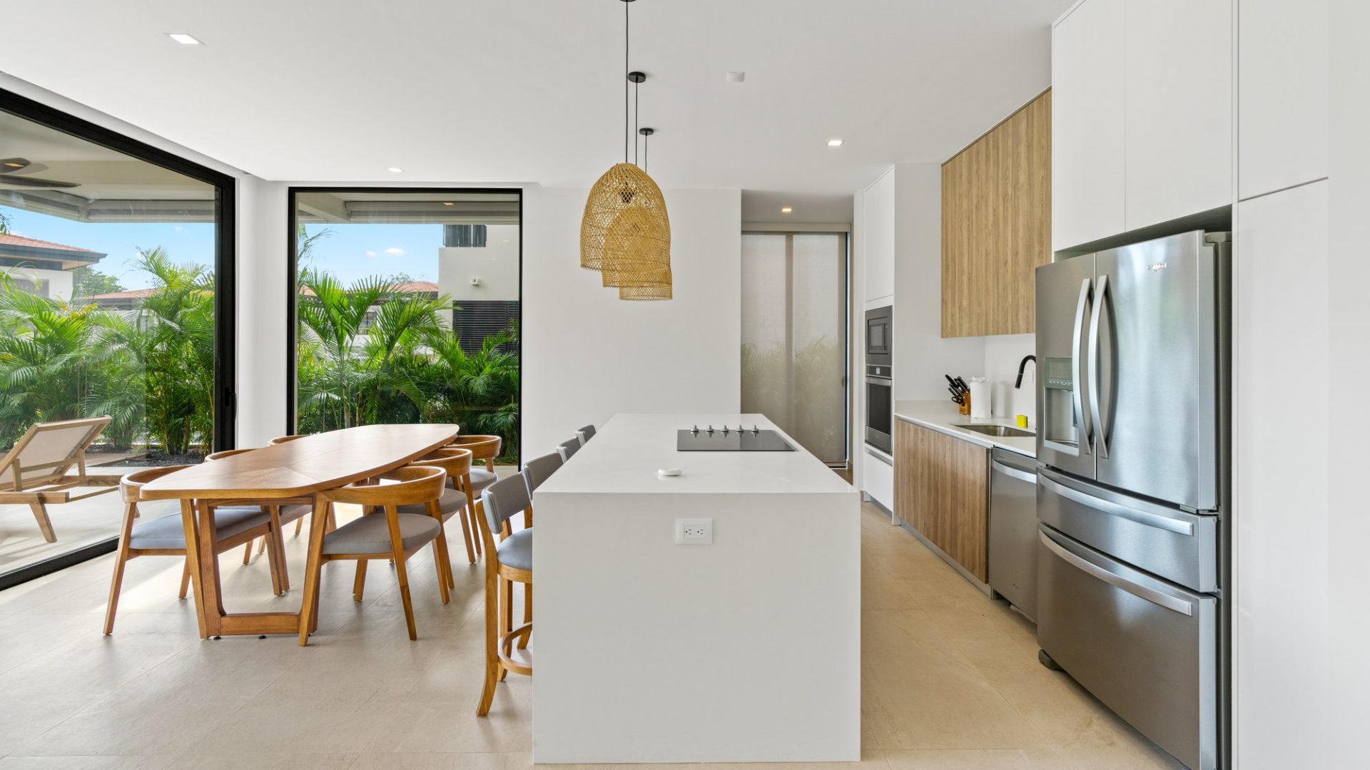 Modern kitchen with dining area and large windows showing lush greenery.