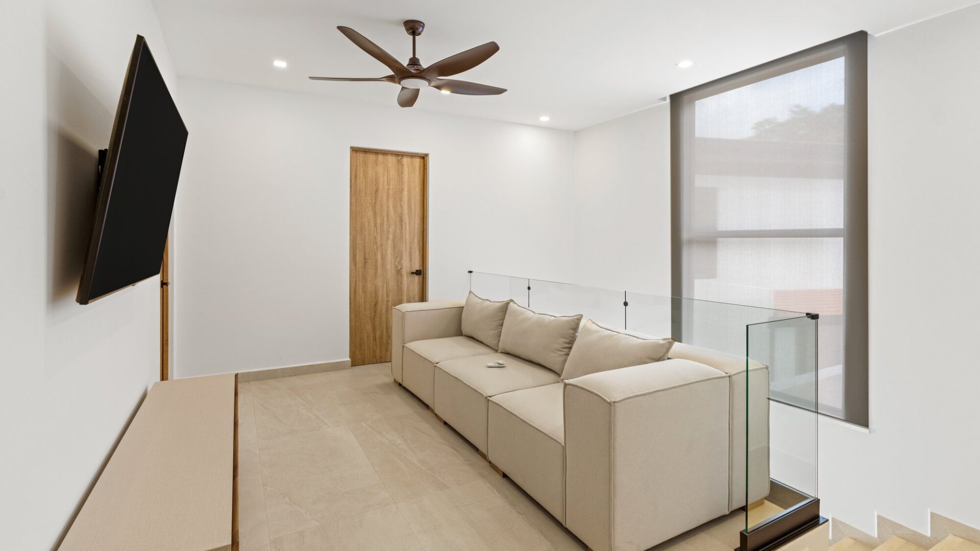 Modern living room with beige sofa, wall-mounted TV, and large window with natural light.
