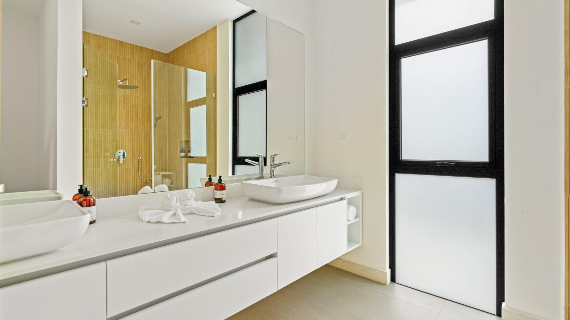 Modern bathroom with white vanity, vessel sinks, and large mirror, featuring a sleek shower area with glass door.
