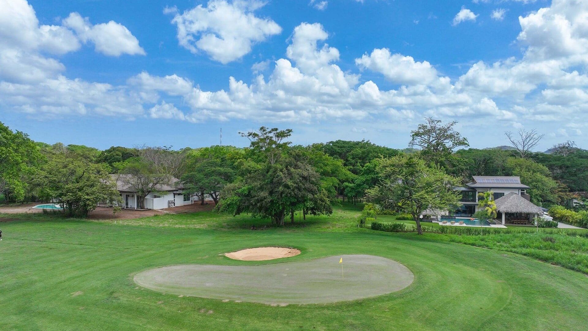 Lush green golf course with sand trap and flag under blue sky with clouds.