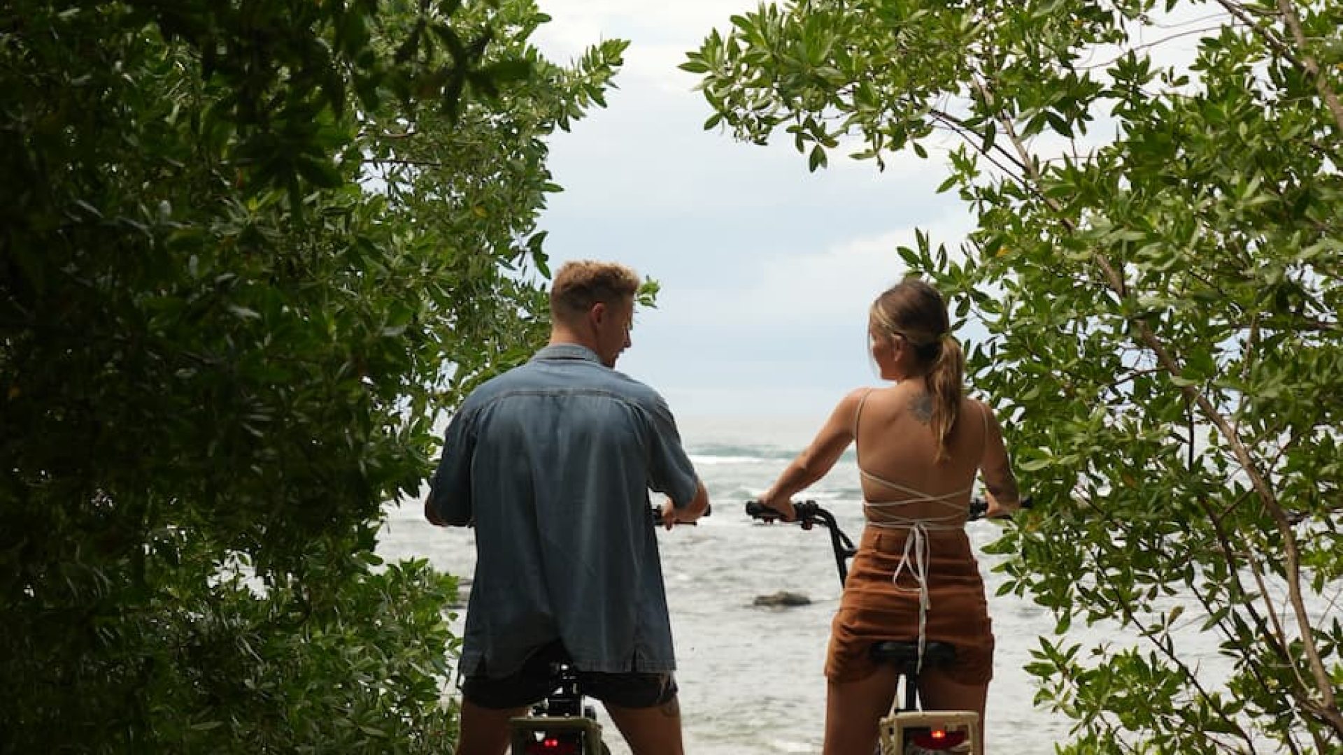 Riding bikes on the beach through lush greenery with ocean view in the background.