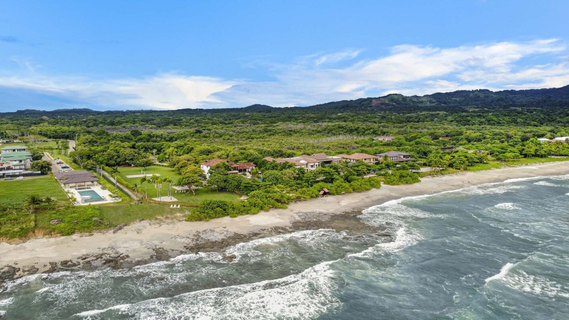 Aerial view of tropical beachfront property with lush greenery, sandy beach, and ocean waves.