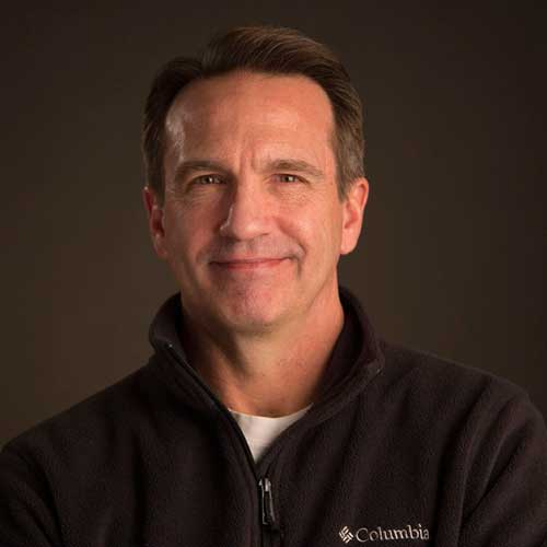 A professional portrait of a smiling man in a black Columbia jacket against a dark background.