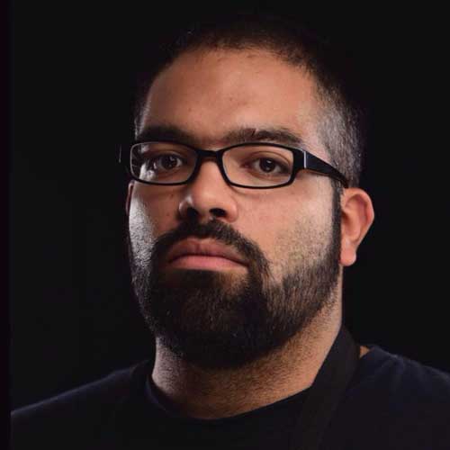 A close-up portrait of a man with glasses and a beard, set against a dark background, emphasizing his facial features and expression in professional lighting.