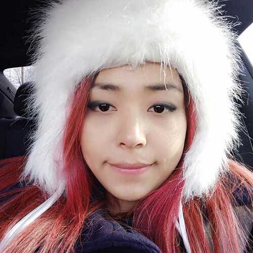 A young woman with pink hair wearing a fluffy white hat, smiling softly, capturing a close-up portrait with natural light.