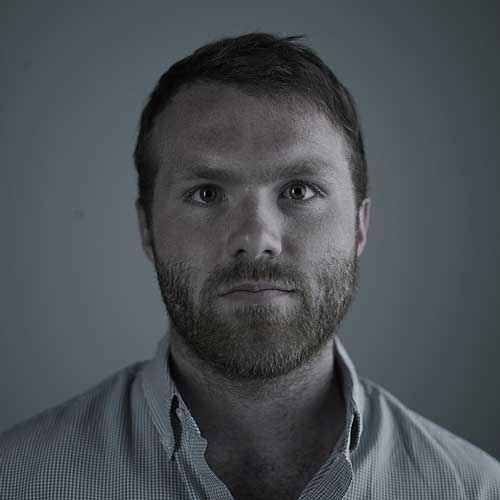 Bearded man with short brown hair wearing a light checkered button-up shirt, facing forward against a plain gray background, in a professional portrait style.