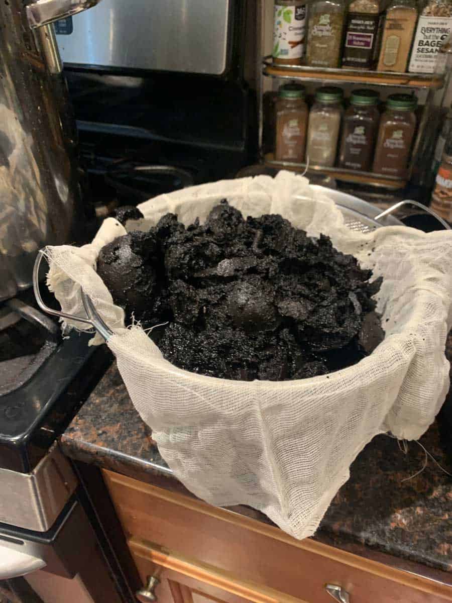 Dark, crumbly activated charcoal in a cheesecloth-lined basket on a kitchen countertop, with spice jars on a shelf in the background.