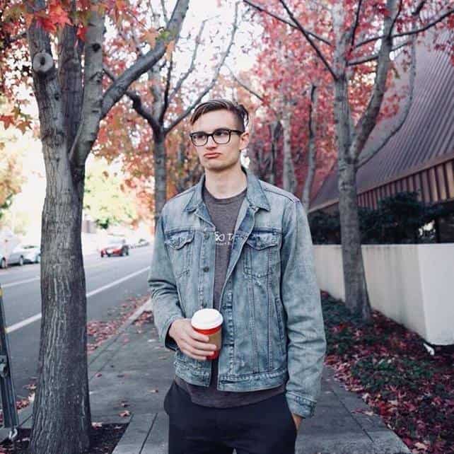 A young man with glasses holding a coffee cup walking along a tree-lined city sidewalk during autumn.