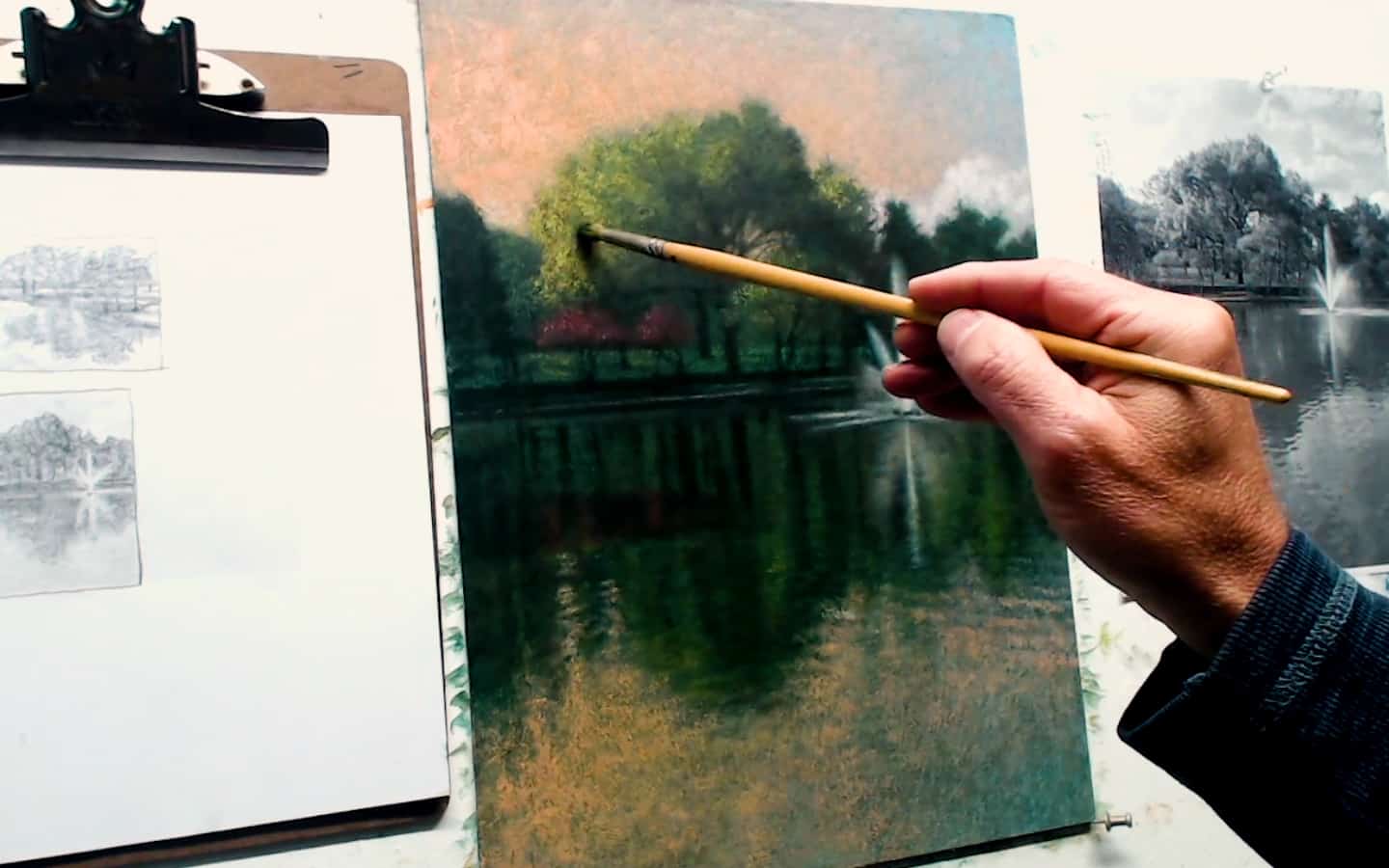 Vibrant landscape painting of a serene lake with lush green trees, created with watercolor techniques on paper at Visual Arts Passage. An artist's hand is seen adding fine details with a brush.