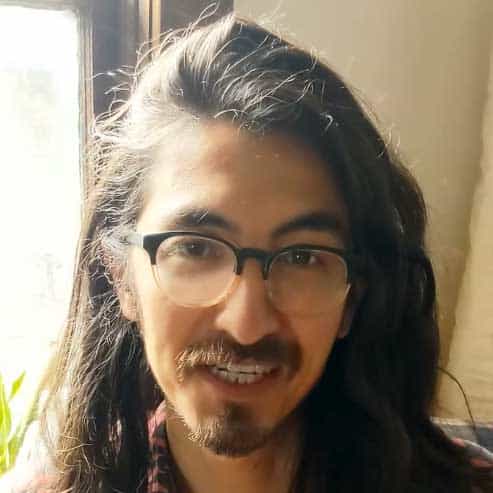 Portrait of a young man with long dark hair, glasses, and a friendly smile indoors, showcasing contemporary art and creative expression.