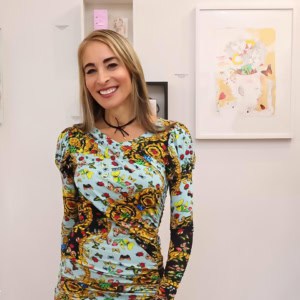 Vibrant woman smiling at art gallery, showcasing colorful floral dress with detailed patterns, standing in front of modern abstract artwork at Visual Arts Passage, emphasizing contemporary art and fashion integration in gallery presentation.
