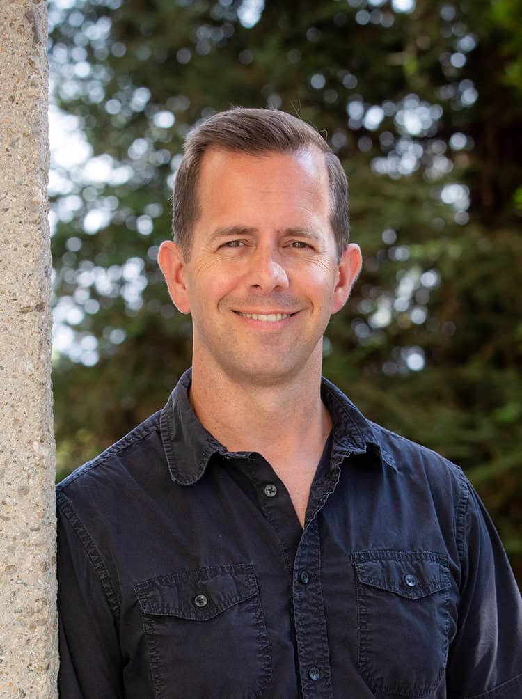 Portrait of a smiling man outdoors with a blurred green foliage background, representing community engagement and personal connection in visual arts.