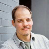 - A professional portrait of a smiling man with a light mustache, wearing a light gray shirt, standing against a gray brick wall, conveying confidence and approachability for Visual Arts Passage.