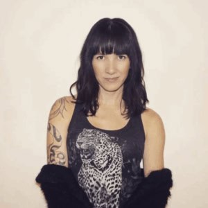 Elegant woman with shoulder-length dark hair, tattoos, and a leopard print sleeveless top, showcasing contemporary art and individual style, against a plain white background.