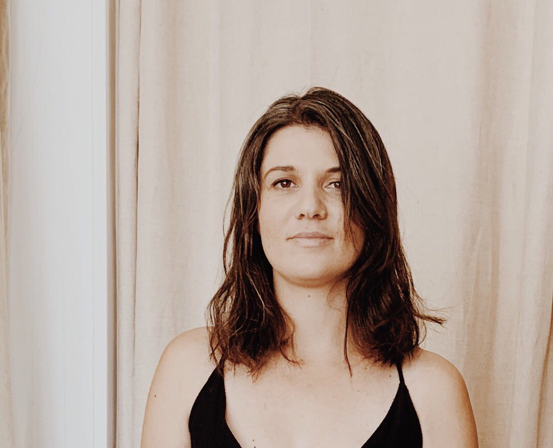 A portrait of a woman with shoulder-length dark hair wearing a black top in front of beige curtains, highlighting contemporary art photography for Visual Arts Passage.