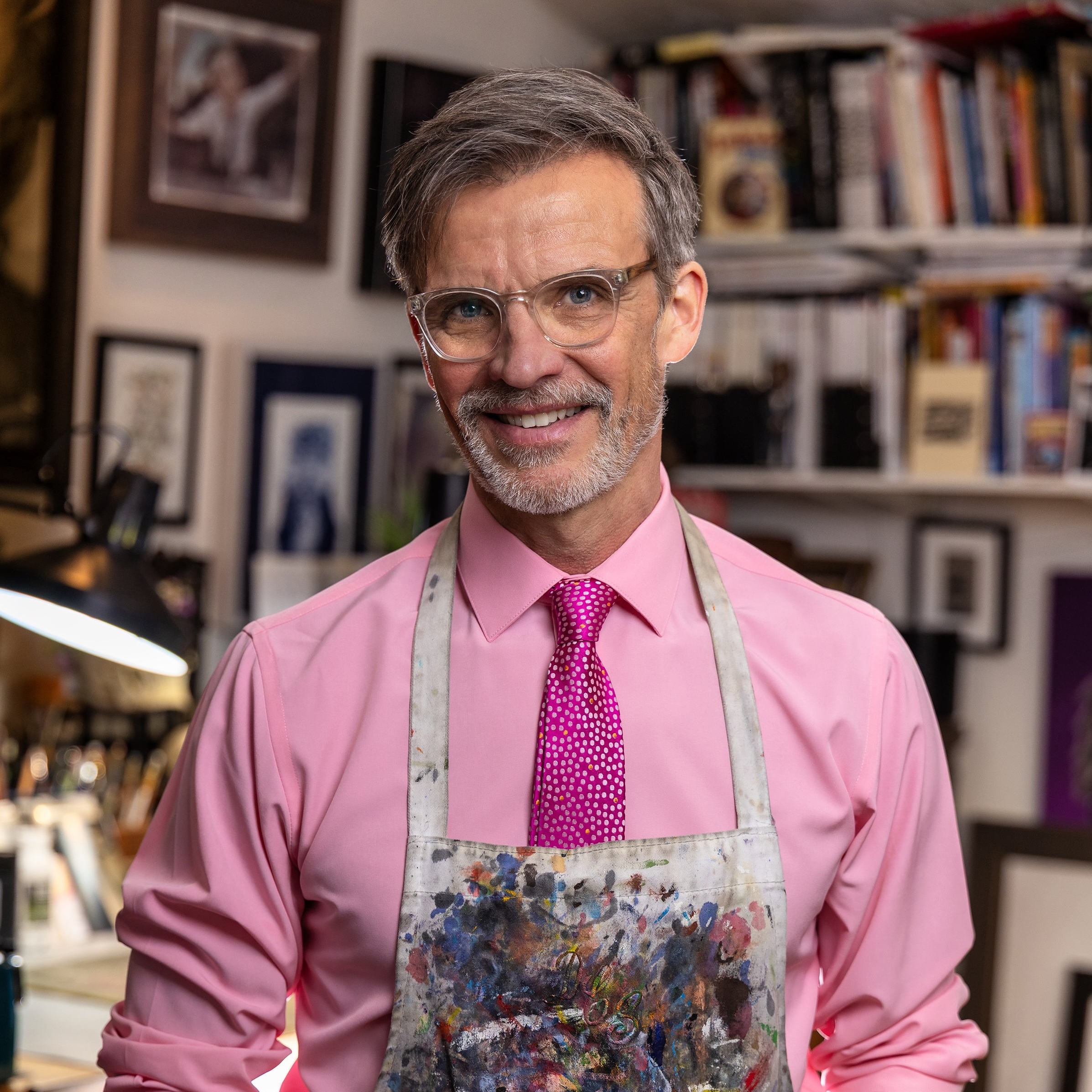 Vibrant middle-aged man smiling in art studio with colorful paintings, art books, and creative workspace background, showcasing passion for visual arts and painting.