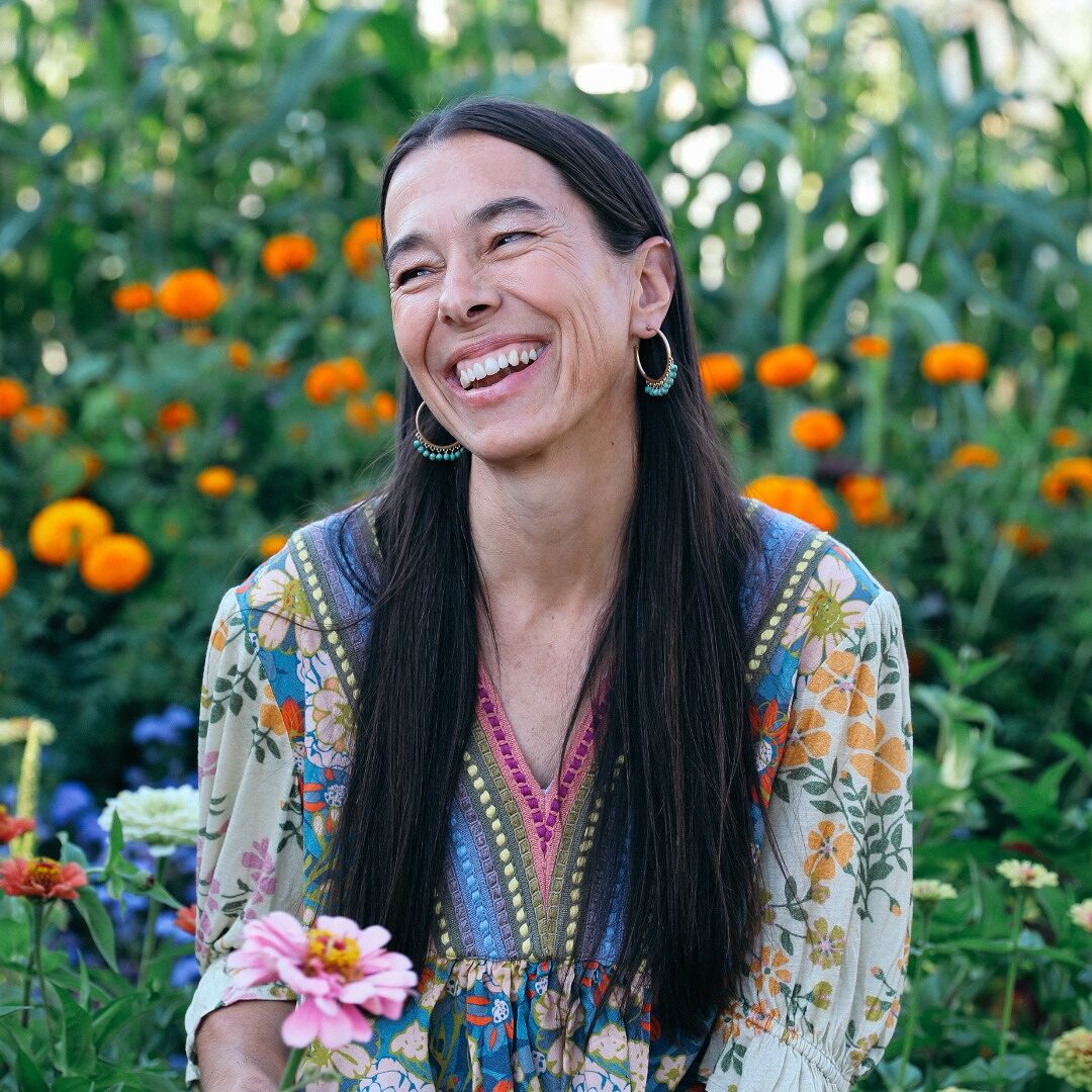 A woman with long dark hair smiling in a vibrant flower garden, wearing colorful bohemian clothing and hoop earrings, surrounded by marigolds and other blooming flowers, exuding joy and natural beauty.