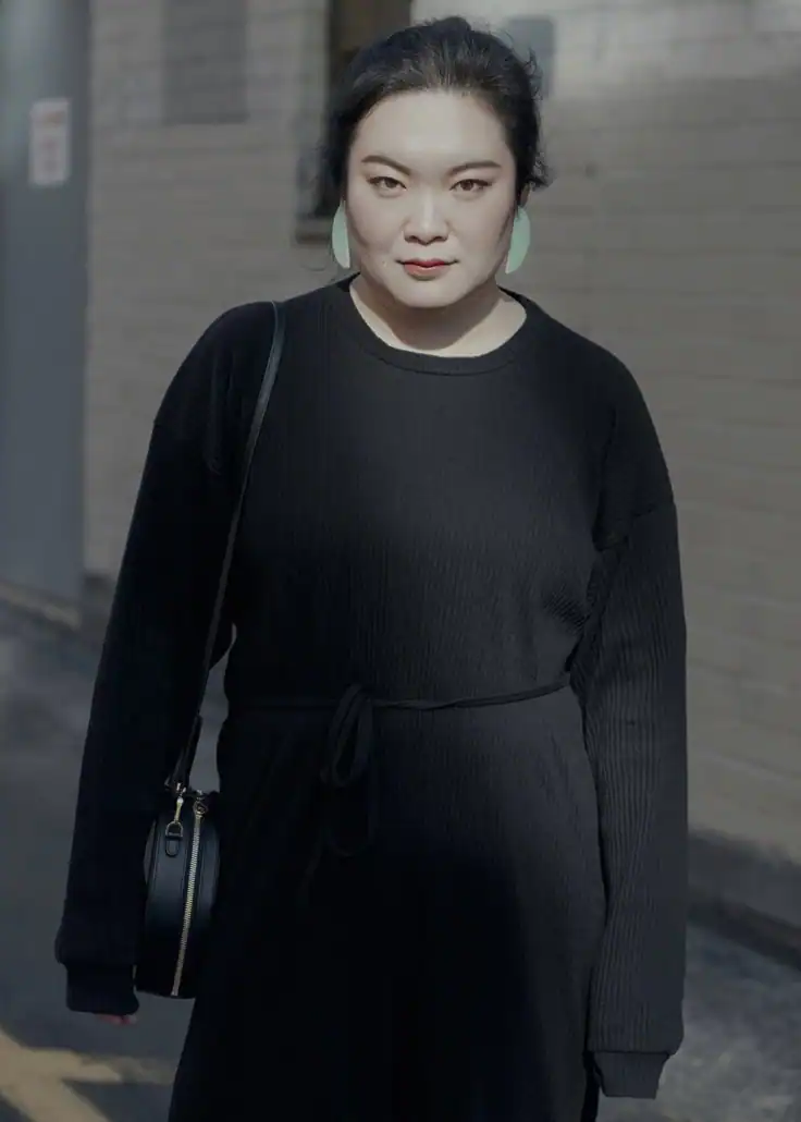 A portrait of an Asian woman wearing a black dress with a black shoulder bag, standing outdoors against a neutral background, showcasing contemporary art and fashion aesthetics.
