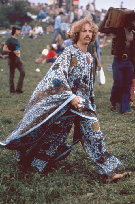 Iconic 1960s hippie at Woodstock music festival wearing colorful, patterned bohemian clothing, holding a guitar, with a crowd of festival-goers in the background.