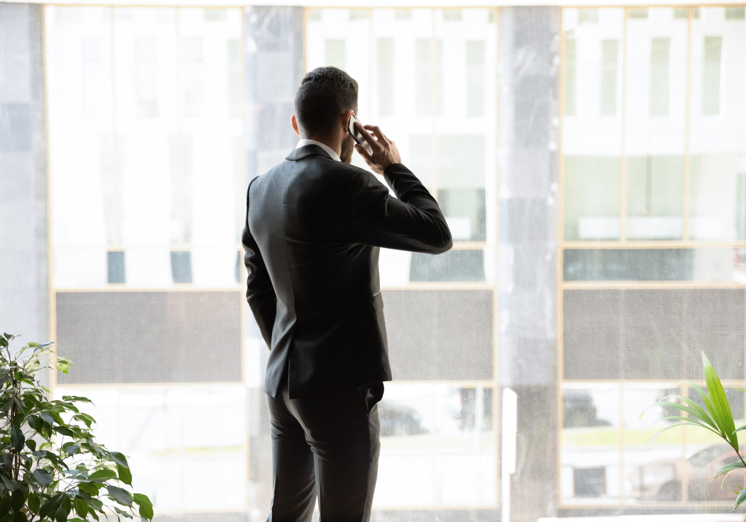 Back view of busy successful businessman in official suit stand near big office window look in distance talking on cellphone, confident man employee speak on smartphone use fast internet connection