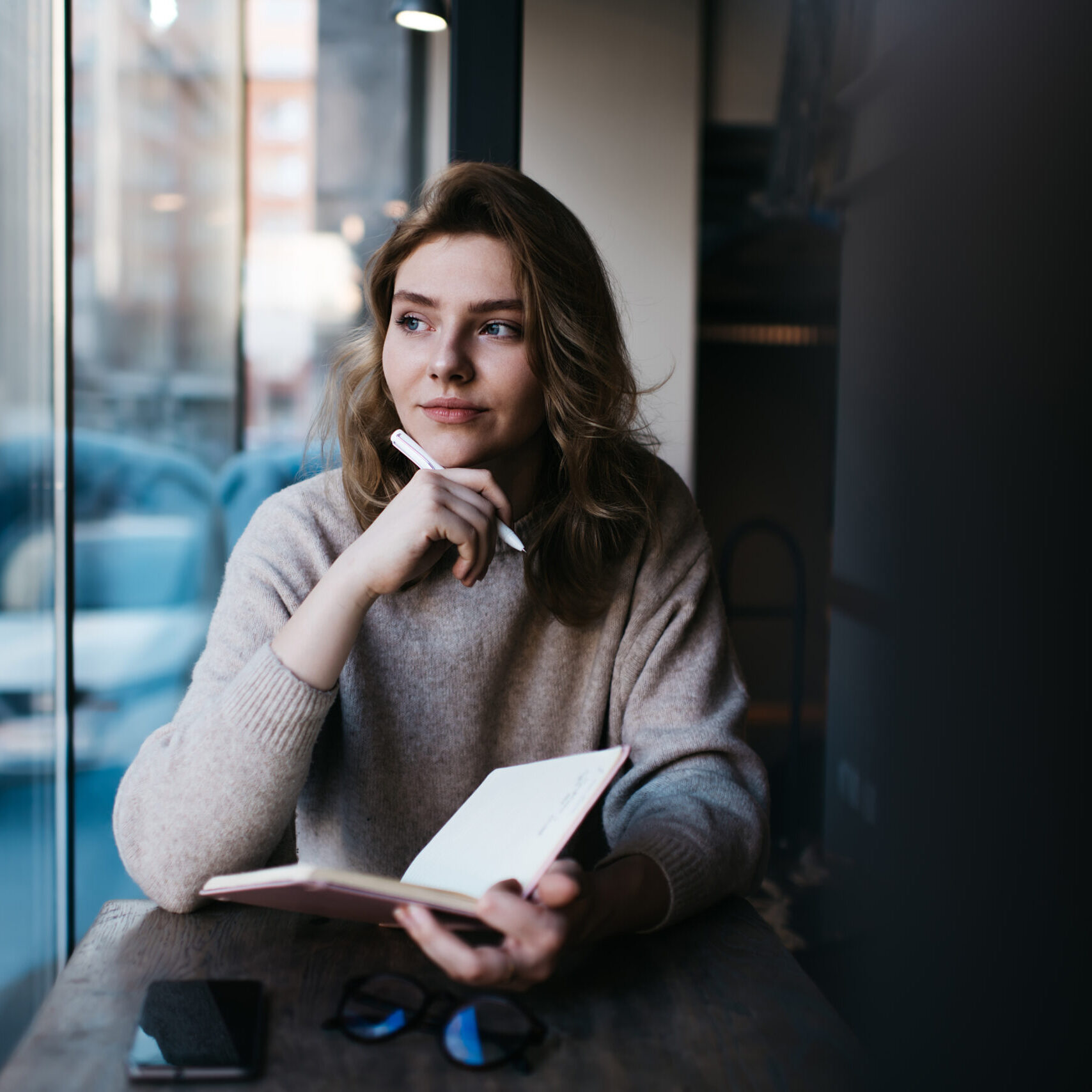 Thoughtful calm female in casual clothes spending time at cafe table with pen and planner and smartphone and eyeglasses thinking about ideas for writing notes while resting near window looking dreamily away