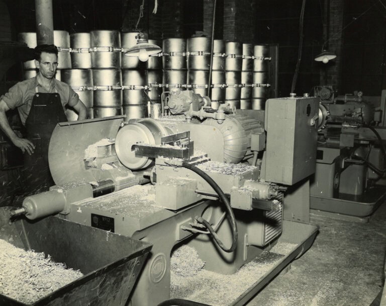 Historical photo of a Wisconsin Aluminum Foundry worker standing in front of sanding equipment, with stacks of cast aluminum pressure cookers in the background and a bin of aluminum shavings in the foreground.